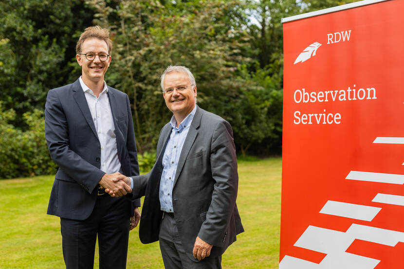 In een tuin op een grasveld staan Daniel Scholz-Stein, CEO van VITRONIC en Zeger Baelde, directeur bedrijfsvoering van de RDW. Beide mannen staan naast een RDW-banner en geven elkaar een hand